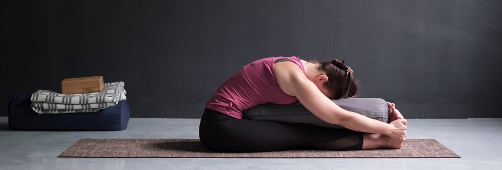 Women stretching forwards over bolster whilst seated with outstretched legs on yoga mat.